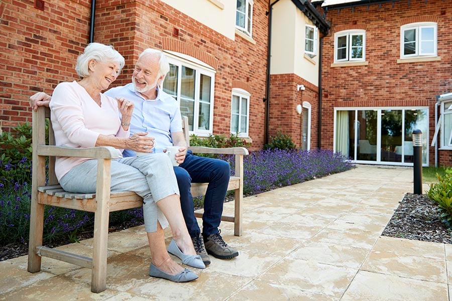 Retired Couple Sitting On Bench With Hot Drink In Assisted Living Facility