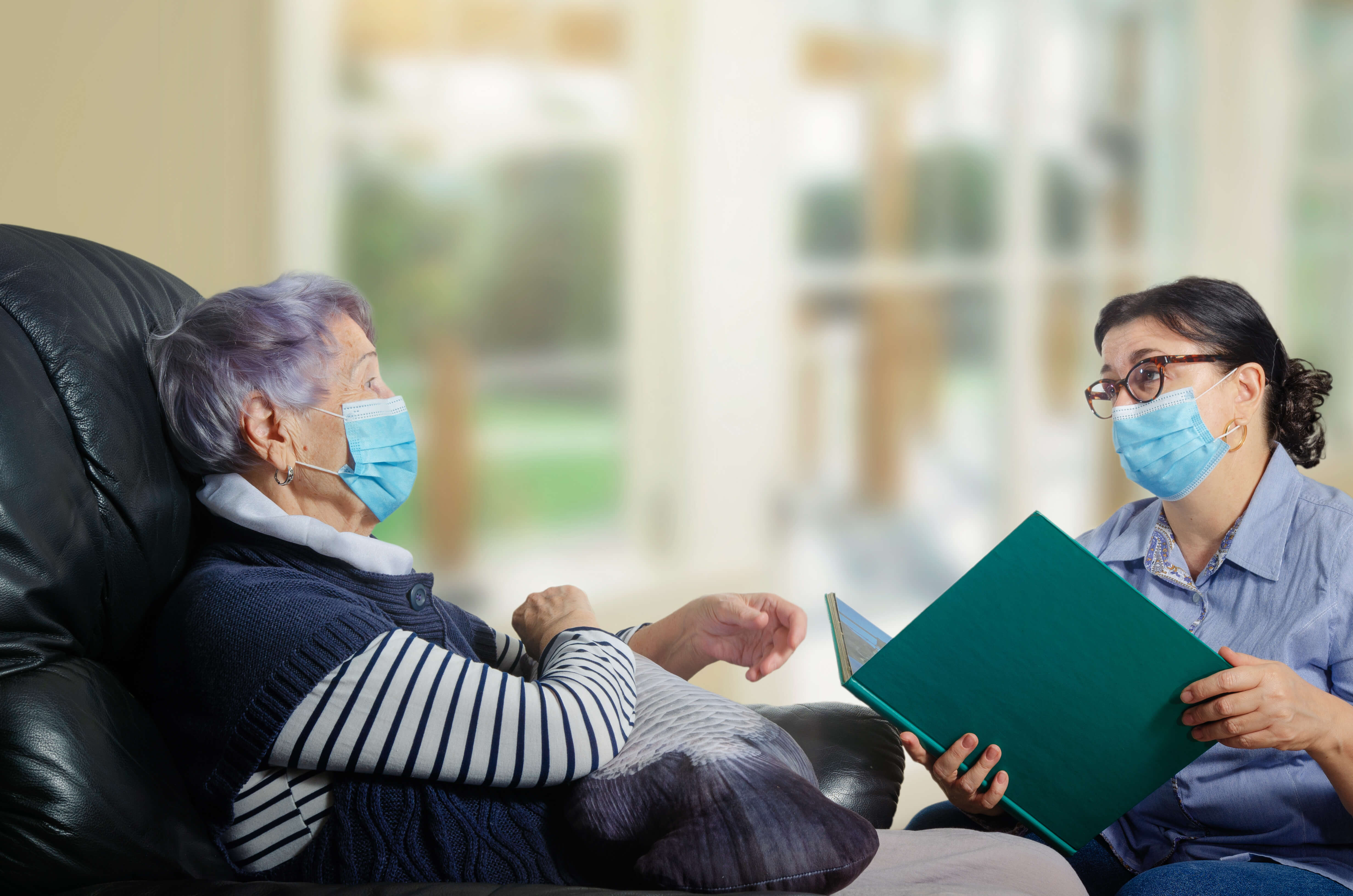 Female adult volunteer wearing a protective mask reading to a senior woman also wearing a mask