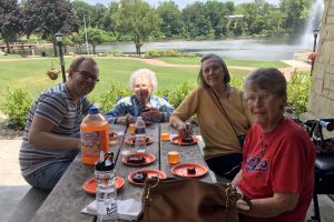Group of female seniors and a gentleman enjoying drinks and dessert outside at a picnic table