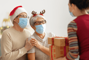 Elderly couple with masks and holiday hats on being greeted by a masked person with presents