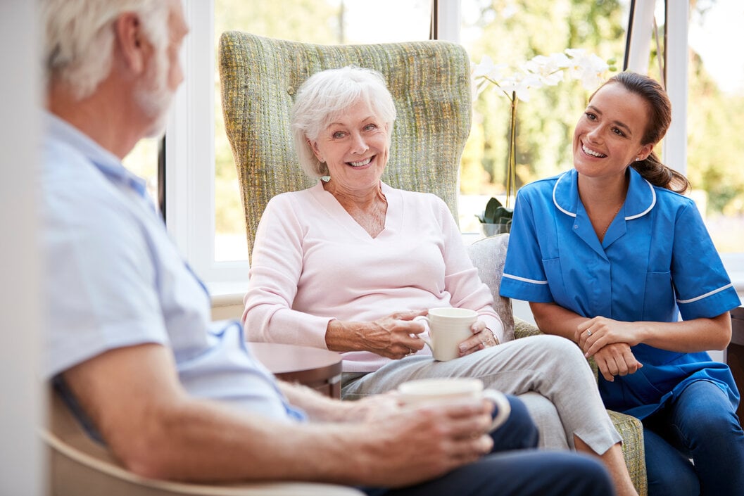 Senior Couple Sitting In Chair And Talking With Nurse In Retirement Home