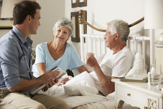 Male doctor talking to an elderly man sitting in bed and his wife next to him