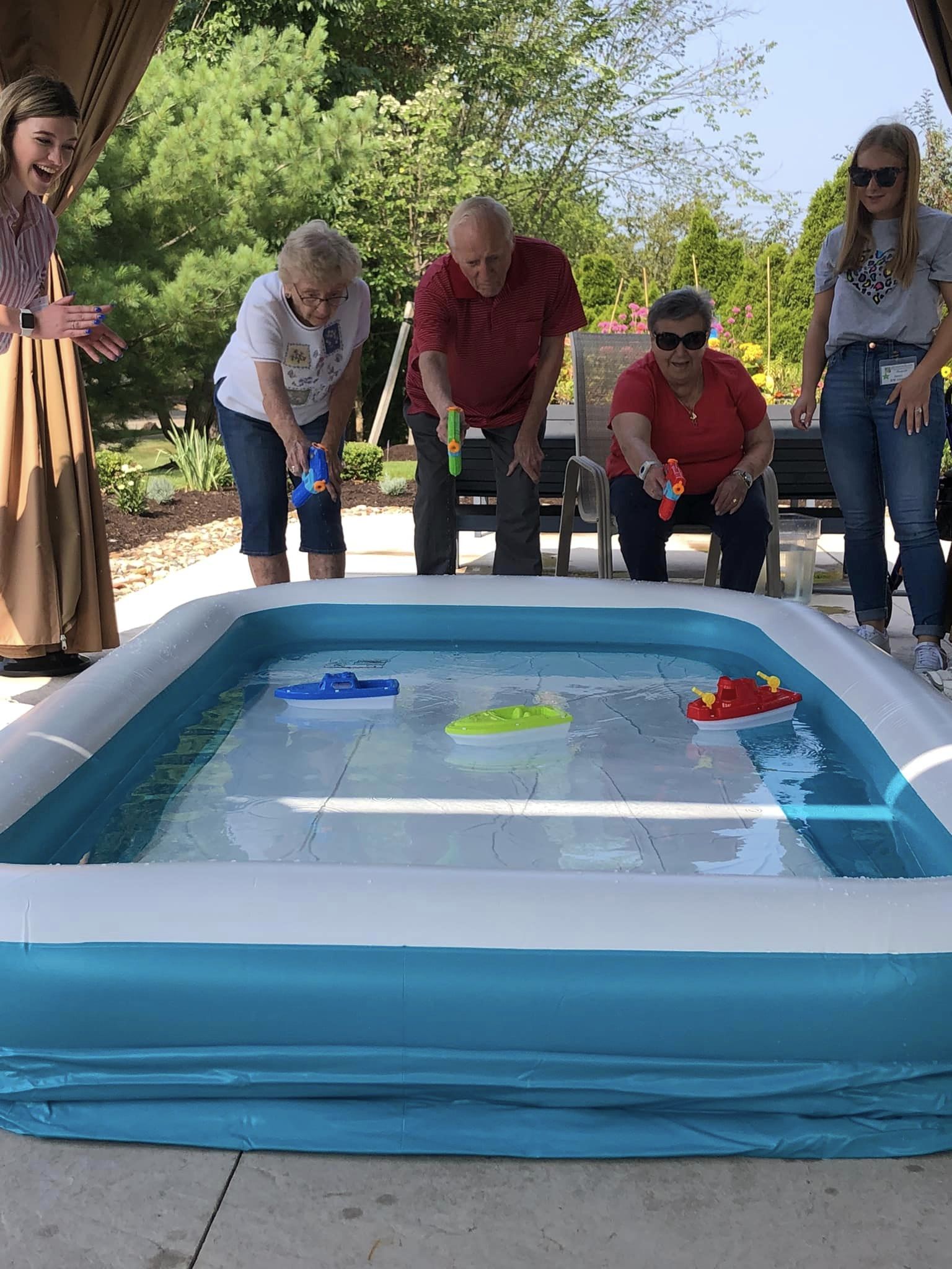 Two senior women and a senior man stand in front of an inflatable pool with three plastic boats in the pool. The three seniors are holding water guns and shooting water at the boats to get them to move.