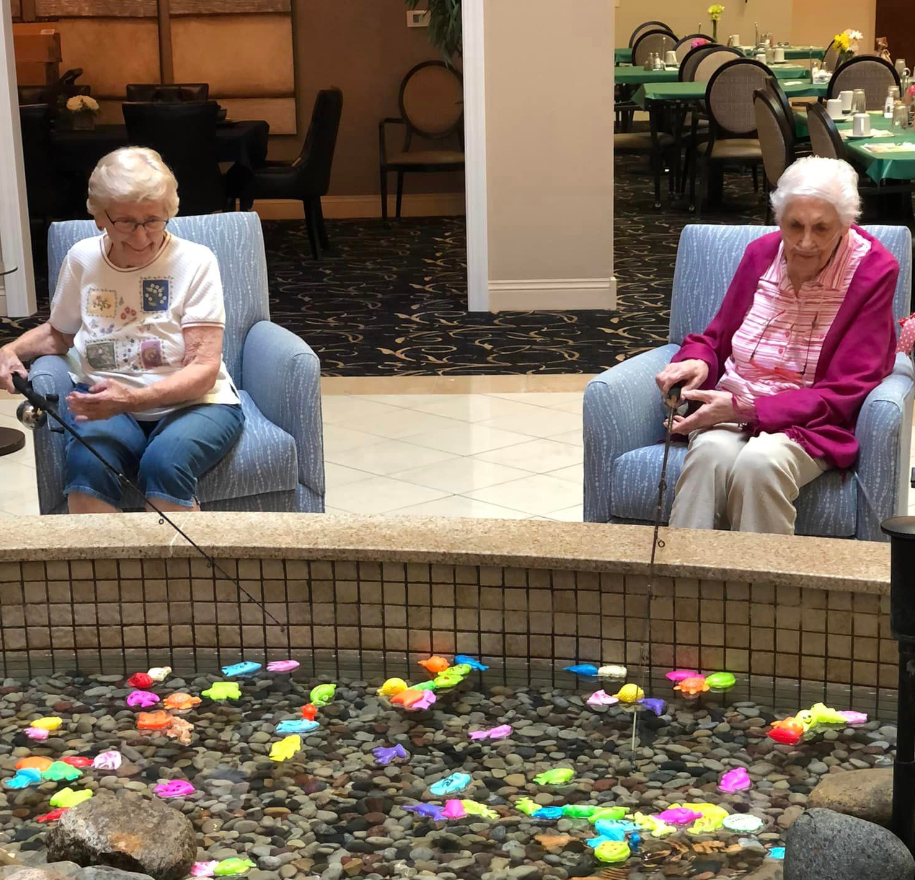 Two senior women sitting in blue chairs holding fishing rods in front of an indoor water fountain. There is a pool of colorful fish in front of them.