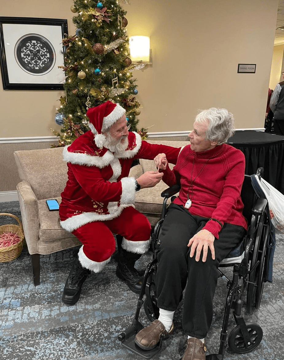 santa clause sits in a brown chair on the left in front of a decorated christmas tree. He looks at a senior woman on the right  wearing a red shirt with a joyful expression on her face-min
