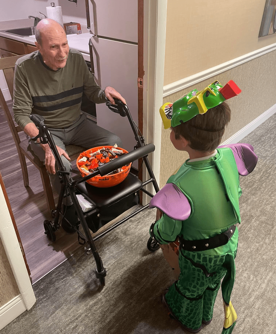 senior man in a green shirt sits in his doorway in front of a large orange bowl of candy with a joyful look on his face. A kid in a turtle costume stands in front of him-min