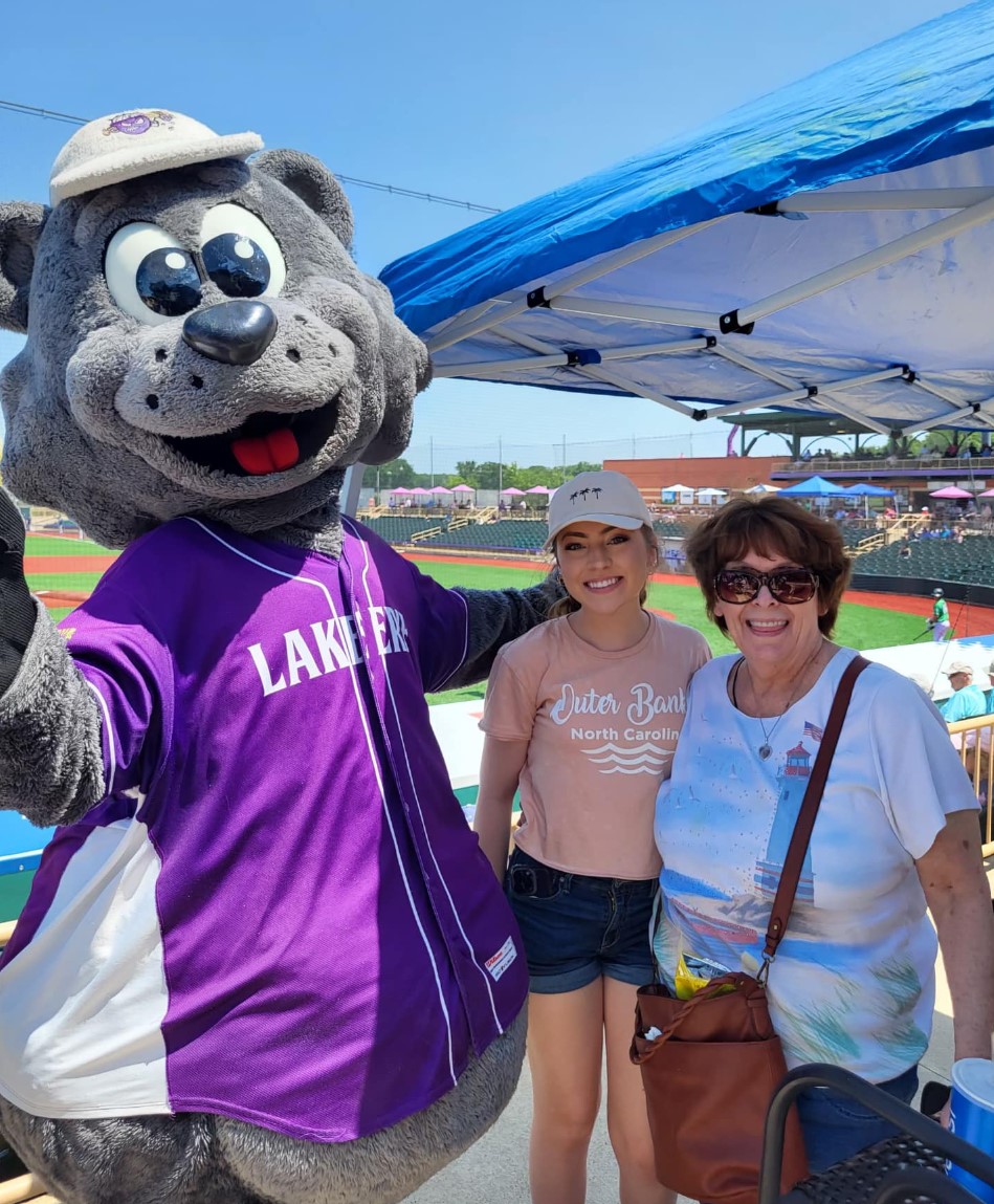 two women standing to the right of a bear mascot in a purple baseball jersey