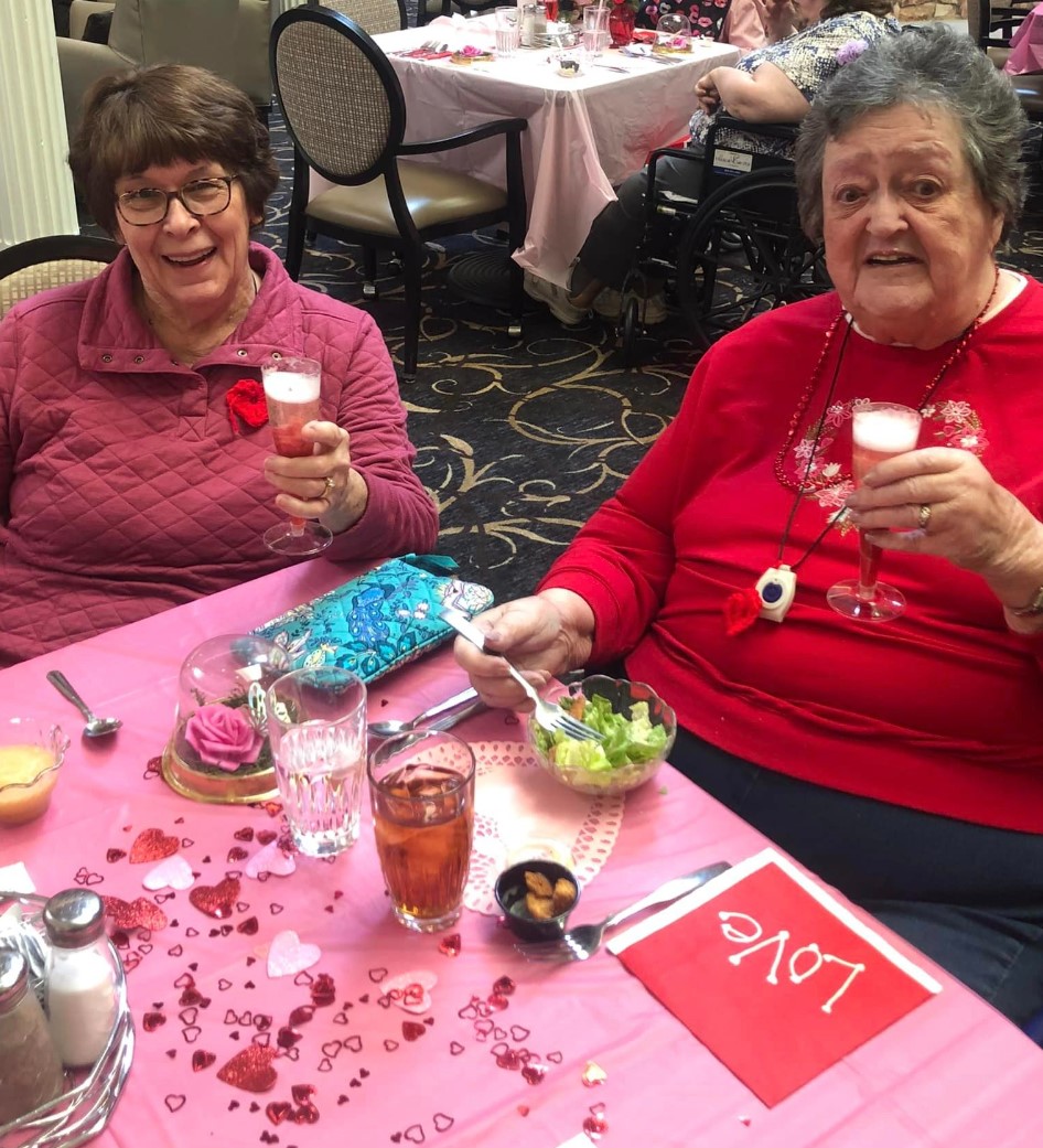 Two senior women sitting at a pink table decorated for Valentine's Day holding a glass of champagne.